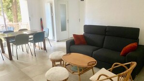 A living room with a black sofa, two wicker chairs, a small table, and a dining area with chairs and a table. - très bel F3 avec balcon (Amélie-les-Bains-Palalda)