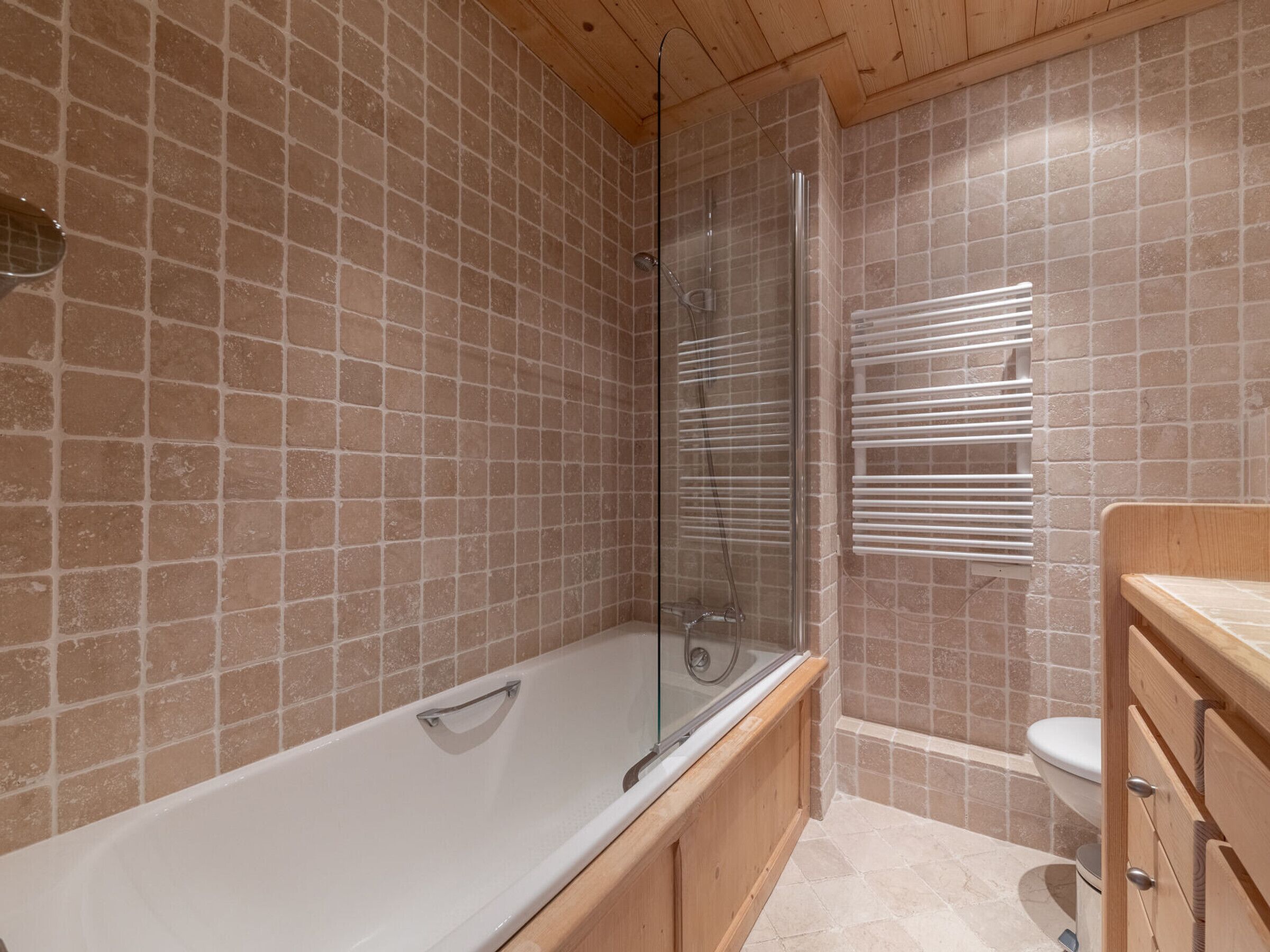 Et badeværelse med et glasafskærmet brusebad, et hvidt badekar og en trækommode med toilet.