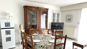 A dining area with a round table, six chairs, a cabinet with glass doors, a television, and a refrigerator.