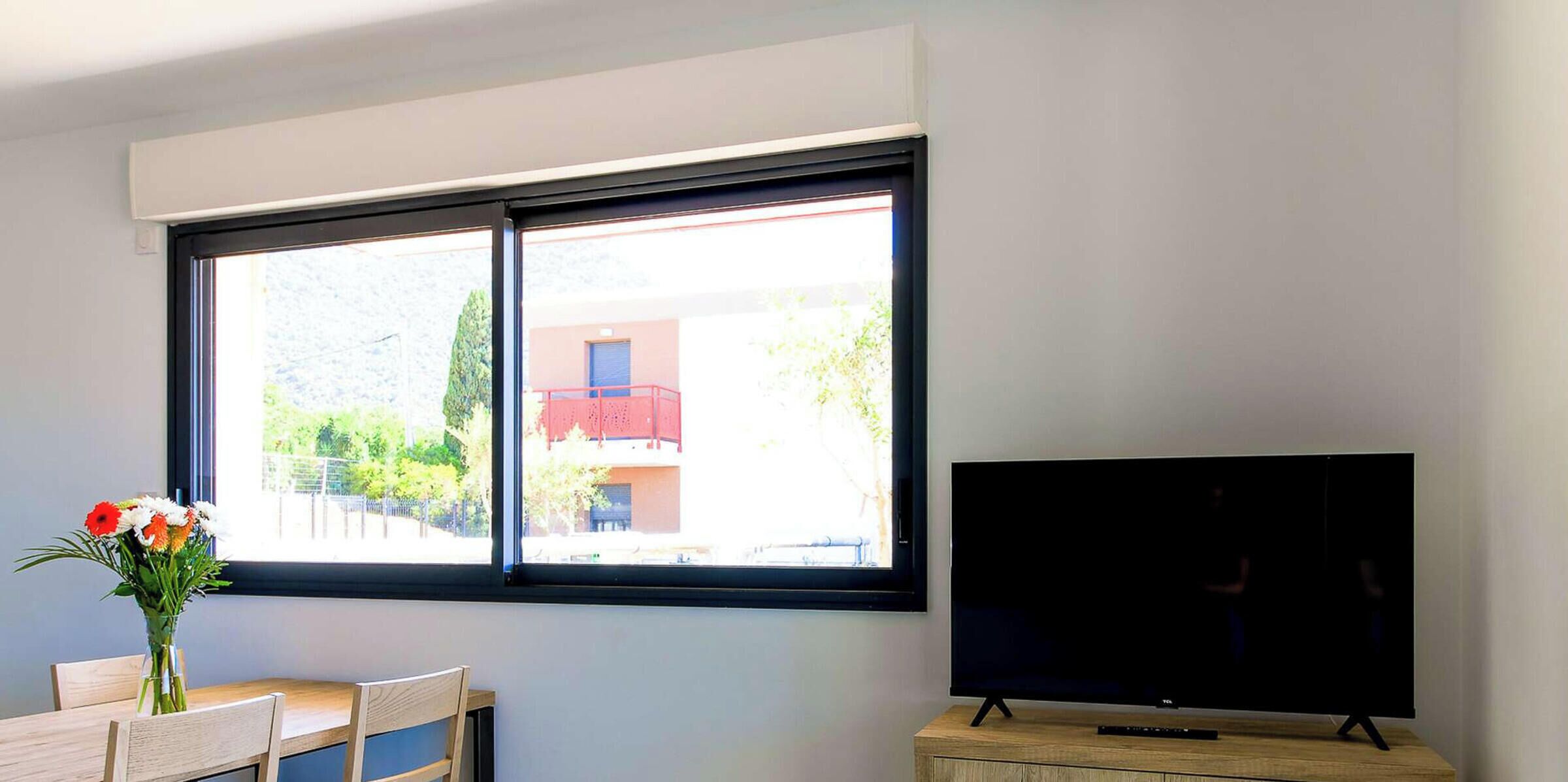 A room with a large window, a flat-screen TV on a wooden stand, a small table with chairs, and a vase with flowers.