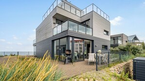 A modern two-story house with a balcony, large sliding glass doors, and a patio area with outdoor furniture.