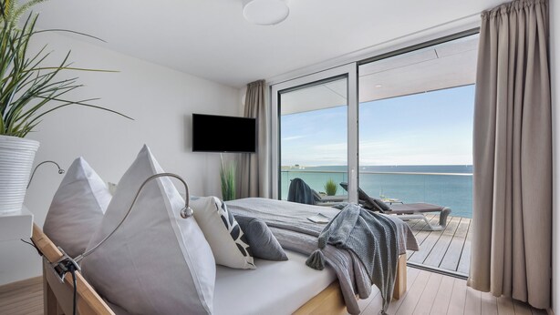 Een moderne slaapkamer met een bed, een televisie, een kamerplant en een balkon met uitzicht op zee.