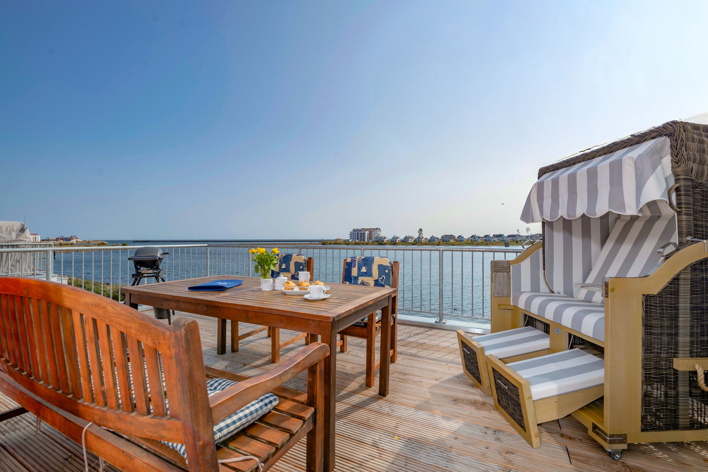 Une terrasse en bois avec une table, des chaises et un coin salon confortable donnant sur une étendue d’eau.