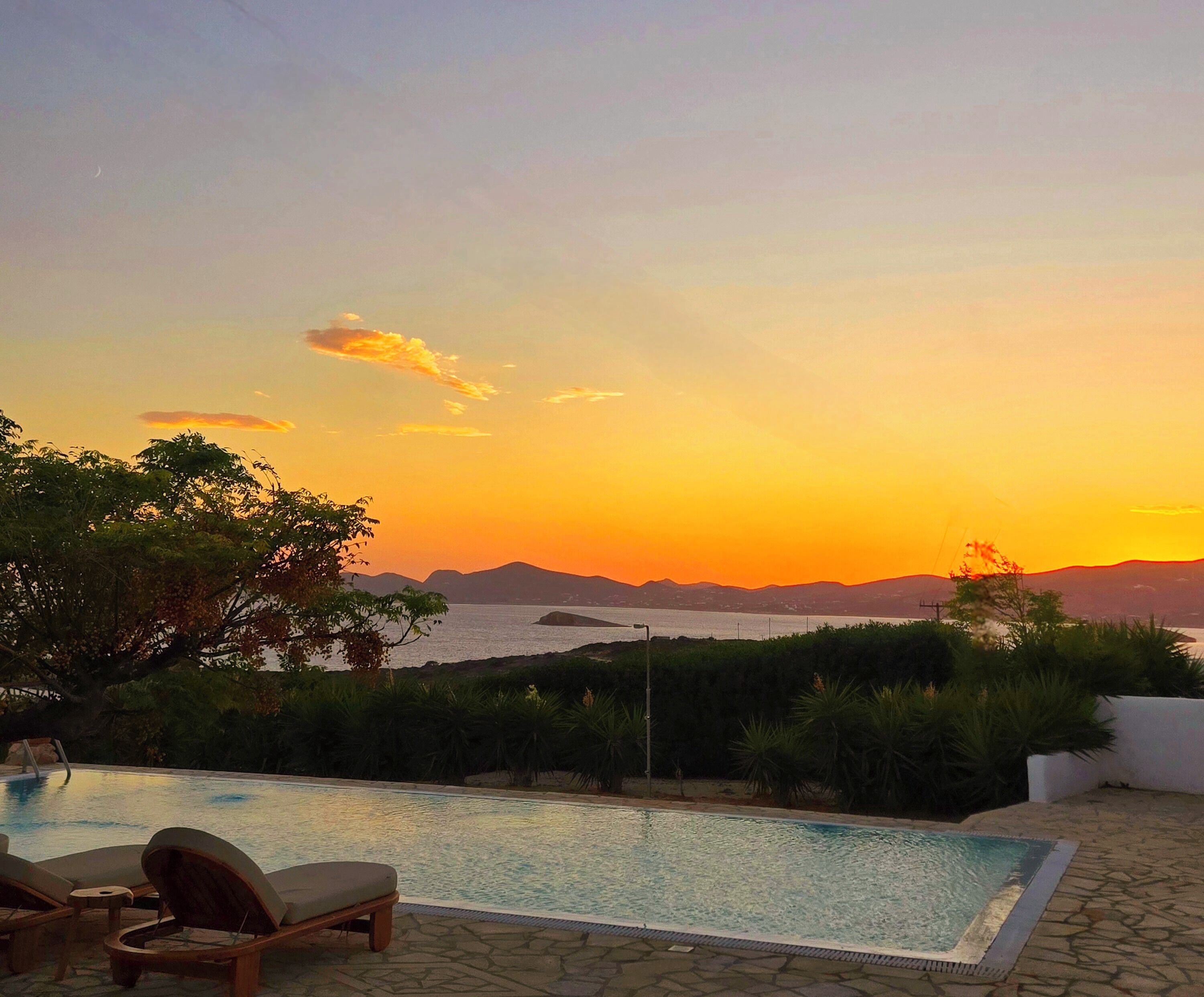 A swimming pool with lounge chairs, surrounded by greenery, overlooking a body of water and distant mountains during sunset.