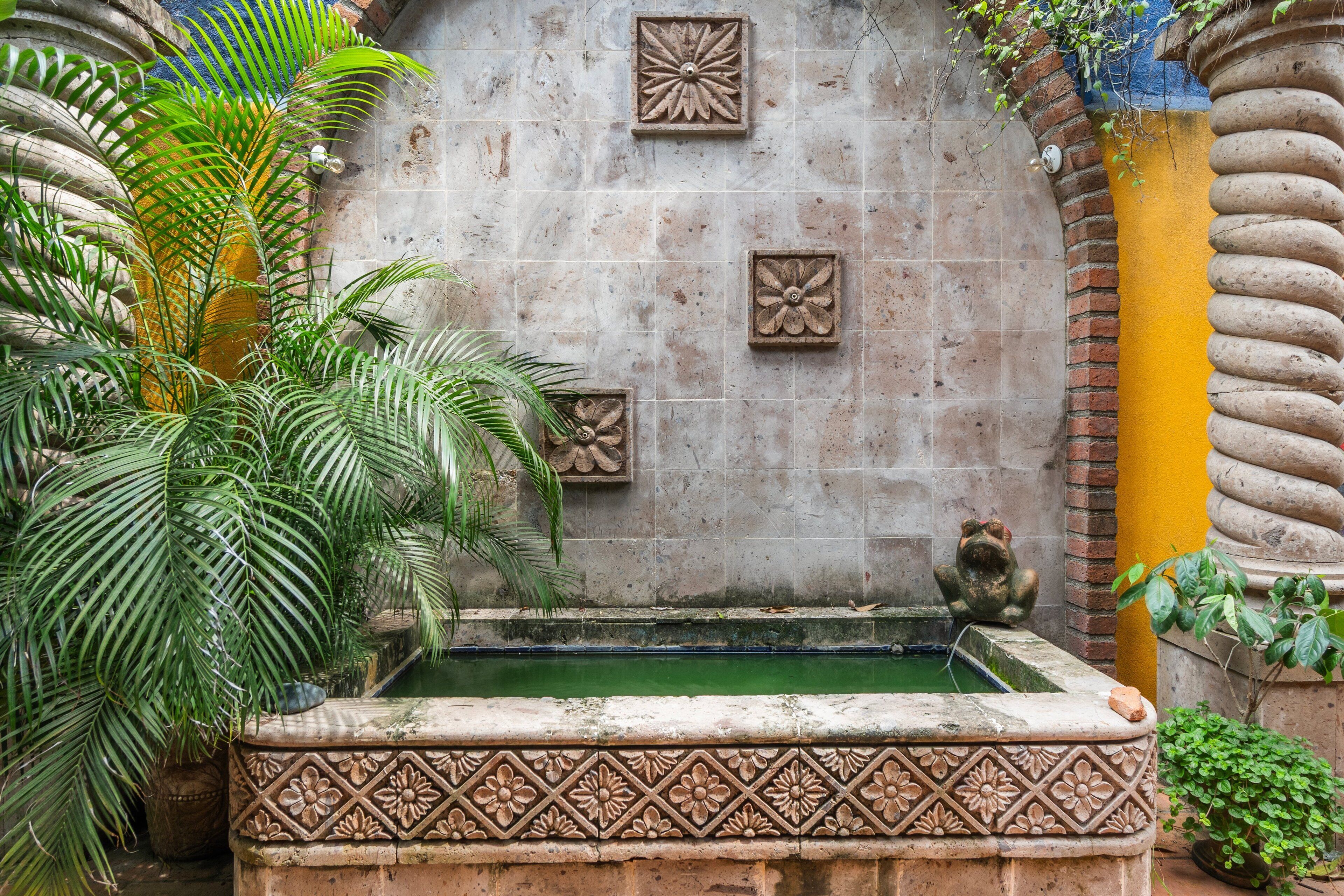 Une cour agrémentée d’une fontaine décorative, d’un mur en pierre orné de motifs floraux et d’une végétation luxuriante.
