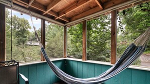 Una hamaca en un porche cubierto con vigas de madera y ventanas amplias que dan a una zona boscosa.