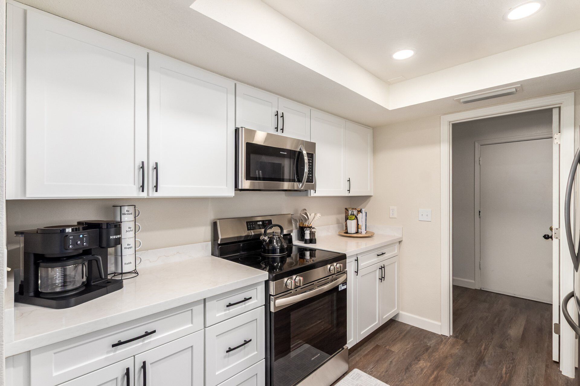 A modern kitchen with white cabinets, a stainless steel microwave, a kettle, and a coffee maker.