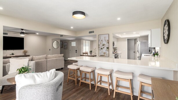 A modern living room with a grey sofa, a coffee table, and a TV. There is a kitchen area with a white countertop and bar stools.