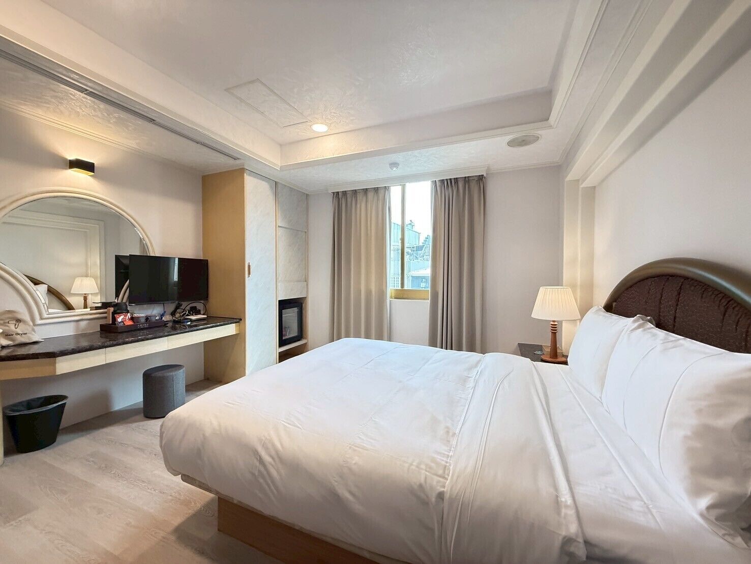 A hotel room with a large bed, a desk with a flat-screen TV, a mirror, and a window with curtains.