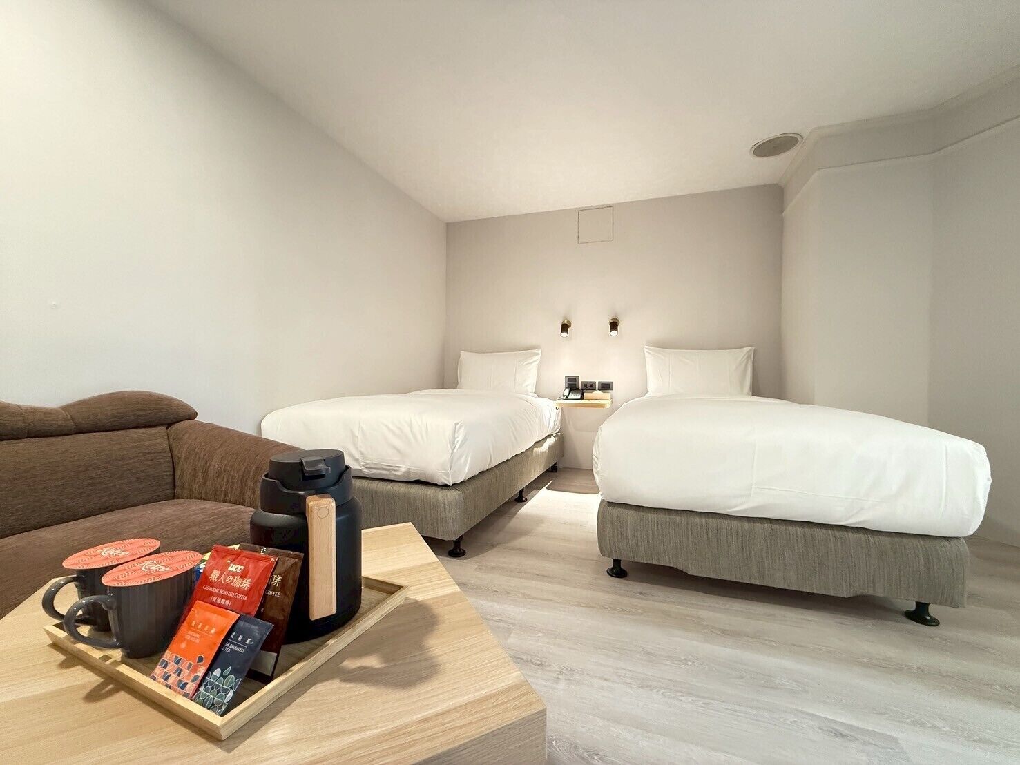 A hotel room with two single beds, a wooden coffee table with coffee packets, and a brown sofa.