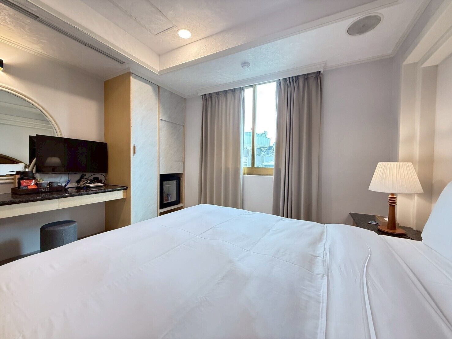 Habitación de hotel con una cama grande, un televisor en un mueble bajo, un escritorio con una computadora y una ventana con cortinas.