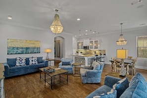 A spacious living room with a blue sofa, matching armchairs, and a wooden coffee table. A kitchen with white cabinets and a stone backsplash is visible in the background. - 4 Bedroom Townhome - Villa Lago at Sandestin Golf & Beach Resort! (Miramar Beach)