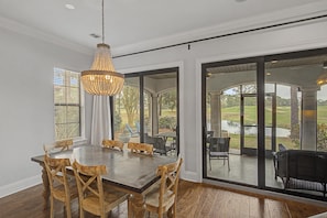 A dining area with a large wooden table and chairs, a chandelier, and large windows offering a view of a garden and outdoor seating. - 4 Bedroom Townhome - Villa Lago at Sandestin Golf & Beach Resort! (Miramar Beach)