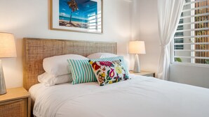 A neatly made bed with white bedding and colorful pillows, a framed beach painting above, and a window with blinds on the right.