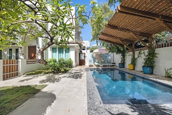 Una piscina con un pérgola de madera, rodeada por un edificio blanco con un patio empedrado.