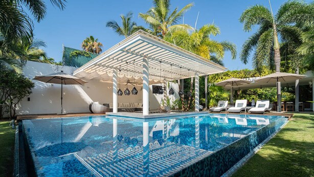 Une piscine agrémentée d’une pergola et de chaises longues, entourée de palmiers et surplombée d’un ciel dégagé.