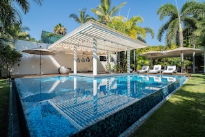 Una piscina con una pérgola y sillas de descanso, rodeada de palmeras y un cielo despejado.
