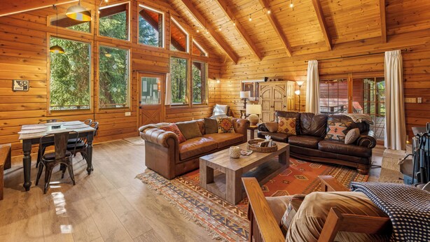 A spacious log cabin interior with a dining area, two sofas, a coffee table, and large windows offering views of the surrounding forest.