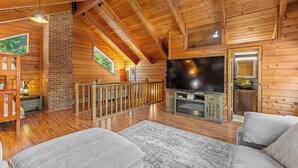 A spacious log cabin interior with a high ceiling, wooden beams, a fireplace, a television, and a cozy seating area.