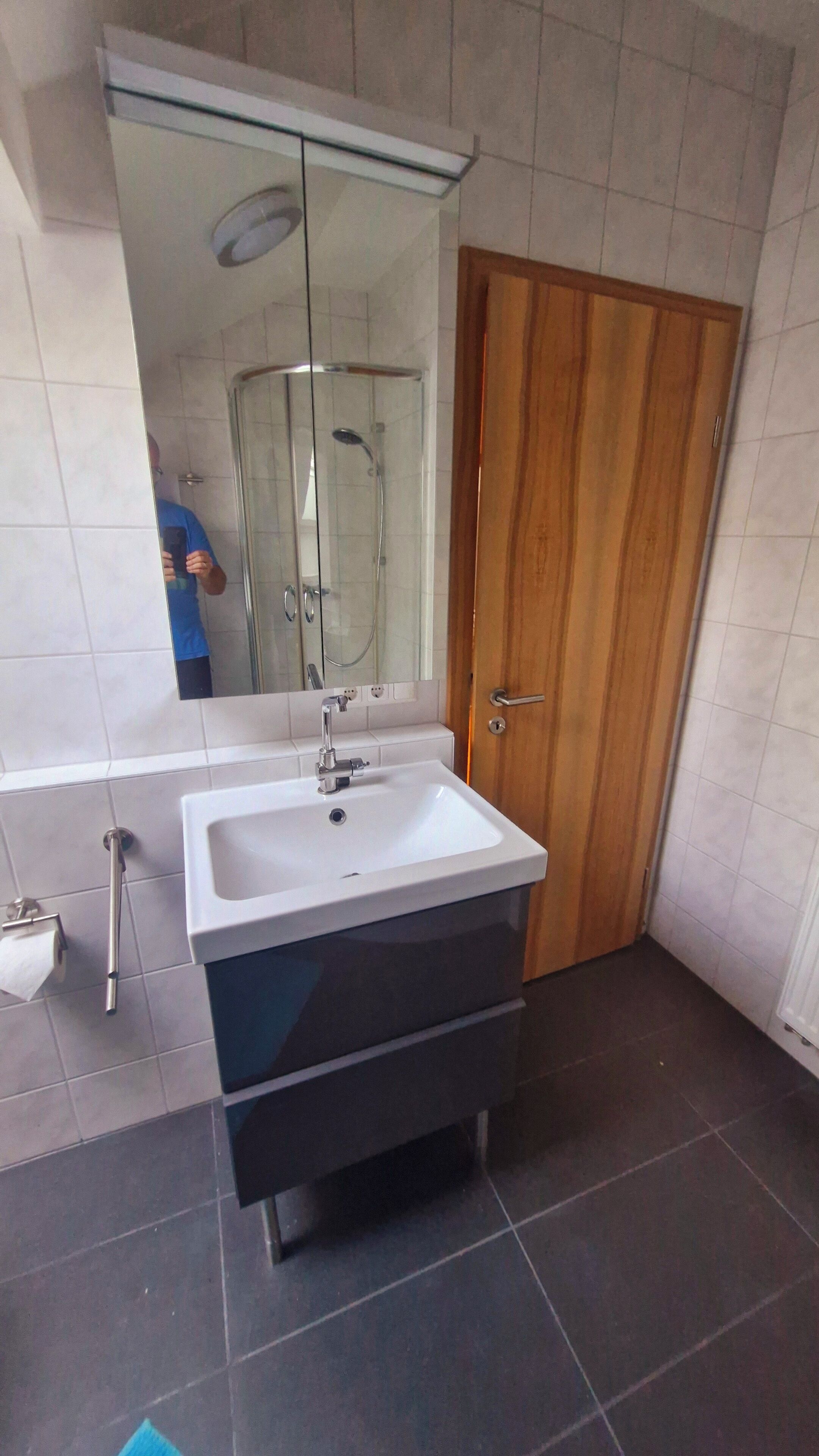 Une salle de bain avec un lavabo blanc, un miroir, une porte en bois et un espace douche.