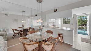 A modern kitchen with a central island, a dining area with wicker chairs and a glass table, and a view of the outdoor pool. - Vacation At Admiral's Bay 148! Fun In The Sun! 2B/2B Condo Walk To Beach! (Fort Myers Beach)