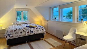 A bedroom with a bed, two bedside lamps, a desk, and a chair. There are two windows providing views of the outdoors.