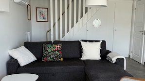 A modern living room with a dark sectional sofa, white armchairs, a white pendant light, a staircase, and a white door.