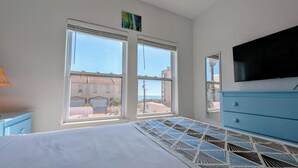 Un dormitorio con cama, televisor montado en la pared y ventana con vista a edificios y palmeras.