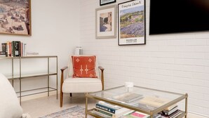 A living room with a glass coffee table, a white armchair with a red cushion, a bookshelf, and framed artwork on the wall.
