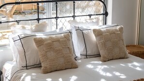 A bed with white linens and beige pillows, a metal headboard, and a view of a stone wall through a window.