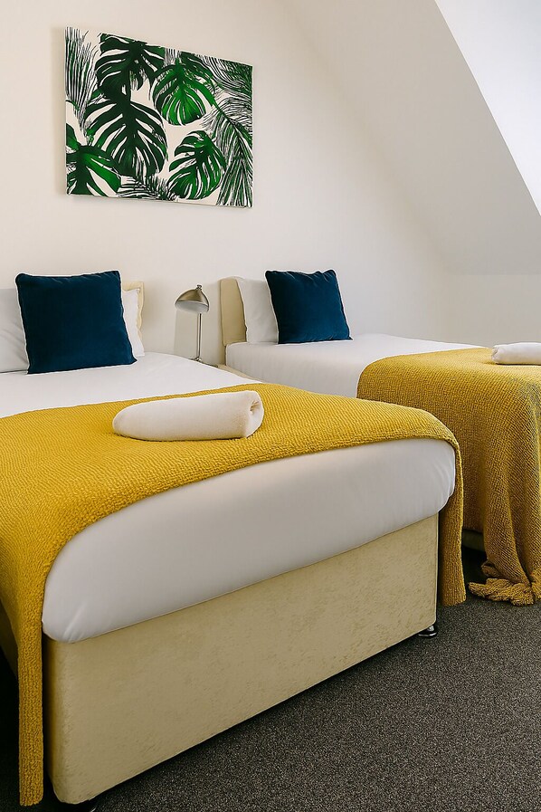A hotel room with two beds, each with white bedding and blue pillows. There is a yellow throw blanket on one bed and a white towel on the other. A large green leaf artwork is on the wall above the beds.