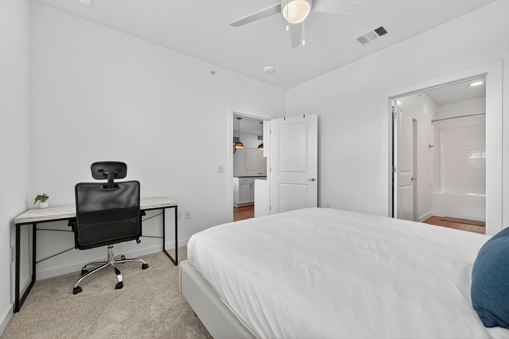 A bedroom with a bed, a desk with a chair, a ceiling fan, and a bathroom with a shower and toilet.