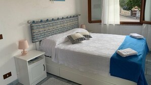 Una cama bien hecha con una manta azul y almohadas, una lámpara de noche y una ventana con cortinas.
