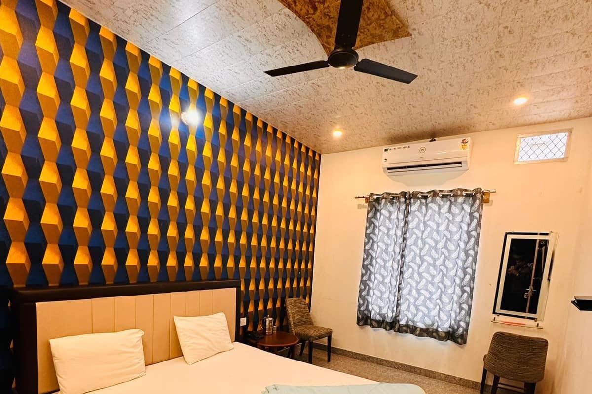 A bedroom with a bed, a ceiling fan, an air conditioning unit, a window with curtains, and a small table with a chair.