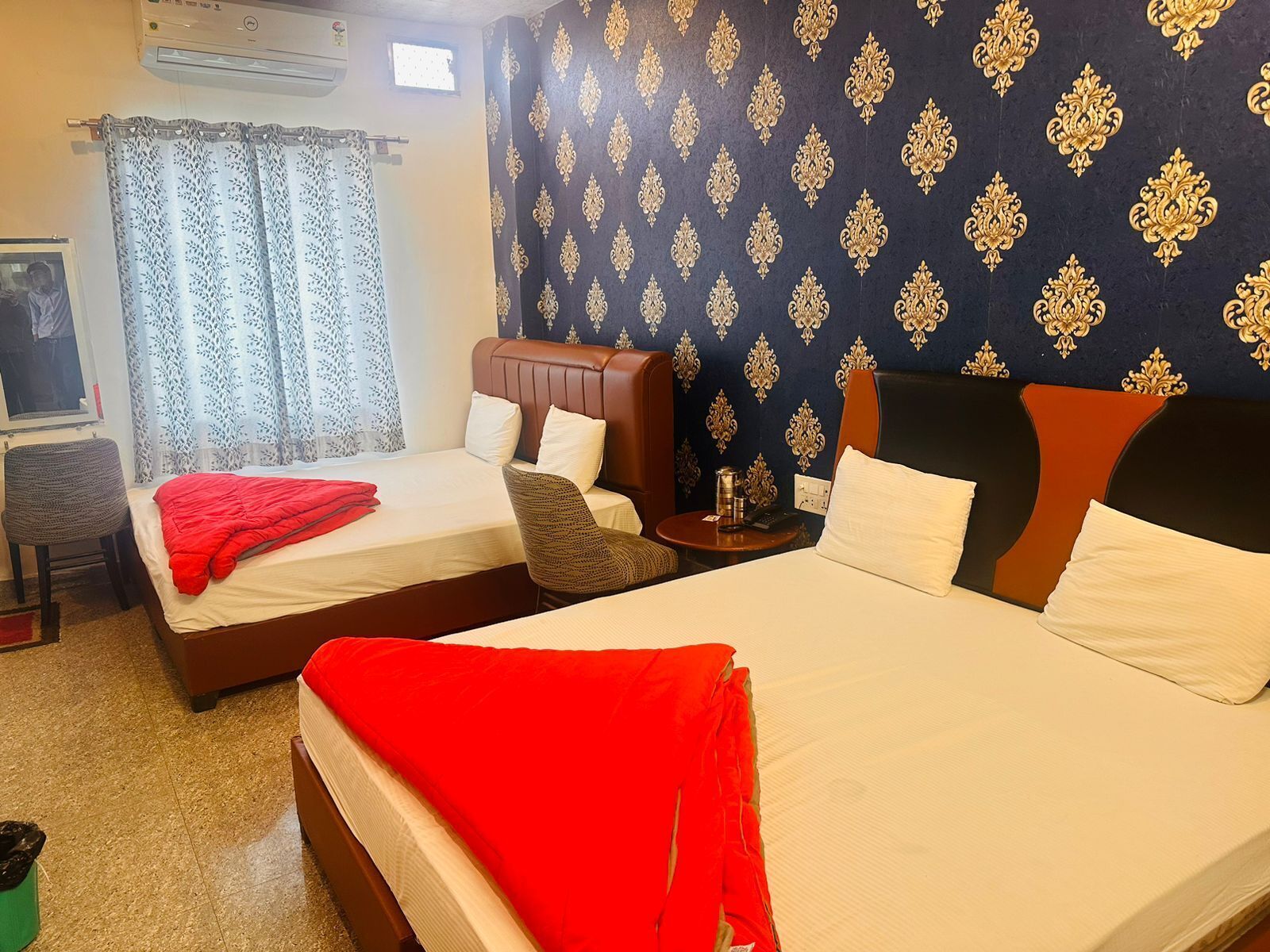 A hotel room with two beds, a small table, a chair, and a window with patterned curtains.