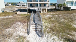 Une propriété située en bord de mer, avec une maison à plusieurs étages dotée d’un balcon, d’une passerelle en bois menant à l’entrée et, en arrière-plan, d’une plage de sable.