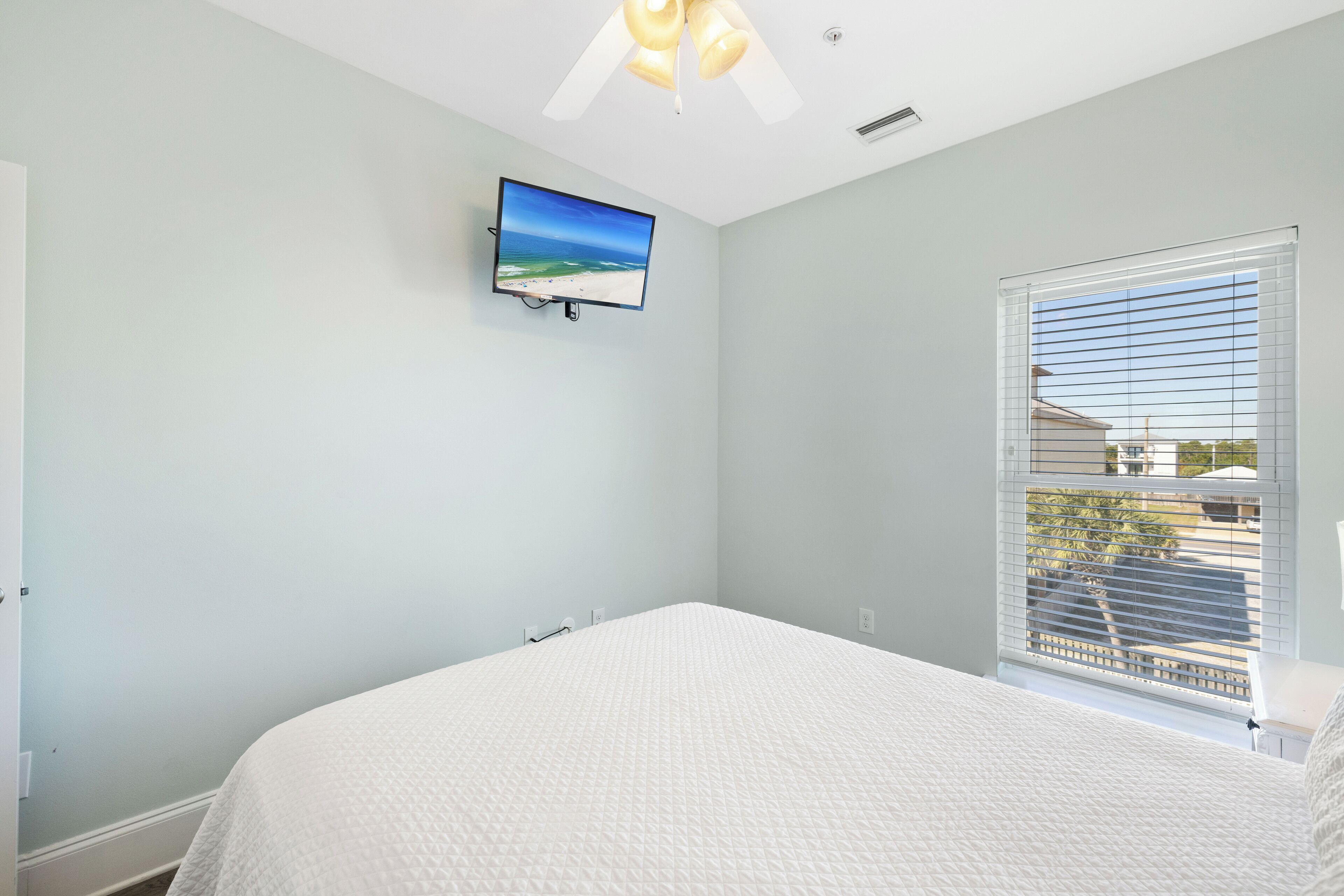 Une chambre avec un grand lit, un ventilateur de plafond, un téléviseur fixé au mur et une fenêtre avec des stores.