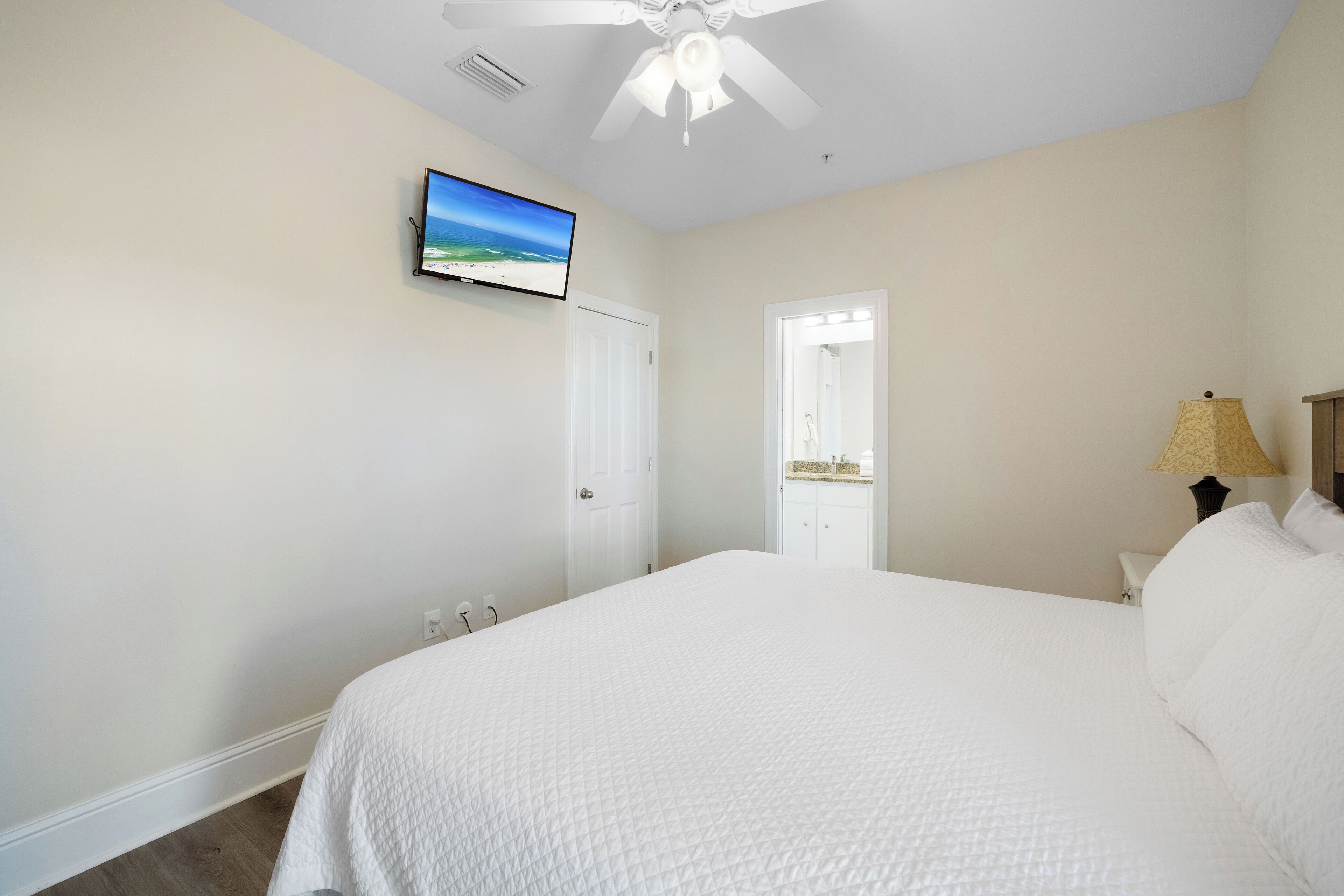 A bedroom with a large bed, a ceiling fan, a TV on the wall, and a door leading to a bathroom.