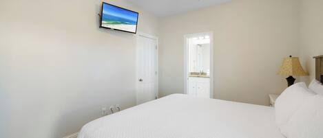 A bedroom with a large bed, a ceiling fan, a TV on the wall, and a door leading to a bathroom.