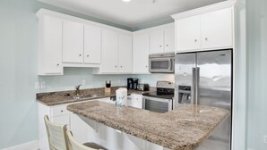 A kitchen with white cabinets, granite countertops, a stainless steel refrigerator, and a built-in oven. - Kiran Terrace Quadplex by Liquid Life (Gulf Shores)