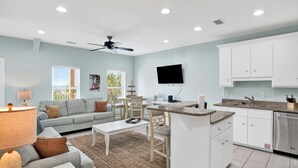 A modern living room with a sofa, coffee table, and dining area. There is a kitchen with white cabinets and a built-in oven. A ceiling fan is visible, and a flat-screen TV is mounted on the wall. - Kiran Terrace Quadplex by Liquid Life (Gulf Shores)