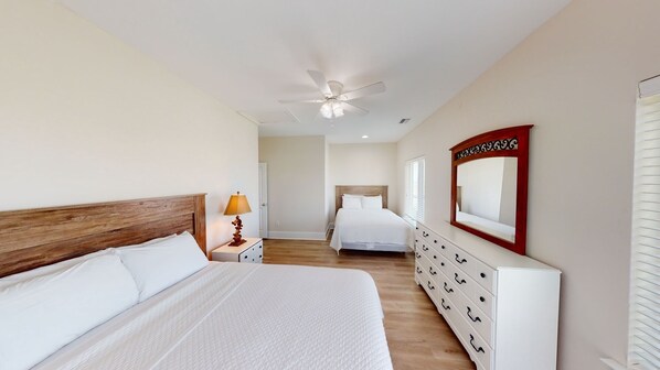 A bedroom with a wooden headboard, a bed with white bedding, a nightstand with a lamp, a dresser with drawers, a ceiling fan, and a large mirror on the wall.