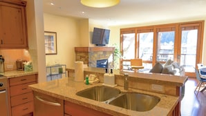 A kitchen with granite countertops, stainless steel sinks, wooden cabinets, a stone fireplace, and a large window with a view of a mountainous landscape.