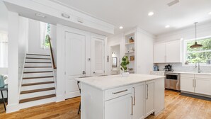 A modern kitchen with white cabinetry, a central island, wooden flooring, and a staircase leading to another room.