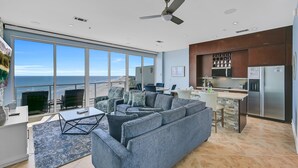 A modern living room with a large gray sofa, a coffee table, and a view of the ocean. - PH, Balcony, Lazy River, I-O Pools, Game Room, Spa, Beach, Luxury 2-2 Penthouse! (Galveston)