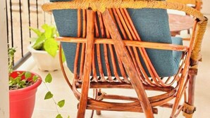 Une chaise en osier avec un coussin bleu, placée sur un balcon doté d’une balustrade, de plantes en pot et d’une autre chaise en arrière-plan.