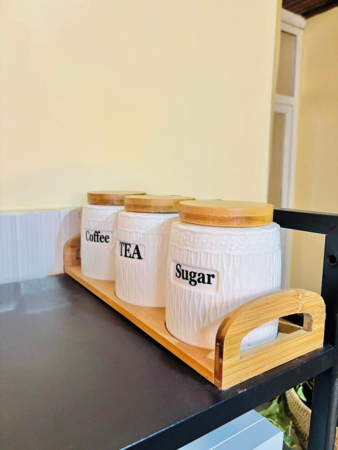 Une étagère en bois avec trois récipients blancs étiquetés « Café », « Thé » et « Sucre ».