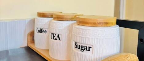 Une étagère en bois avec trois récipients blancs étiquetés « Café », « Thé » et « Sucre ».