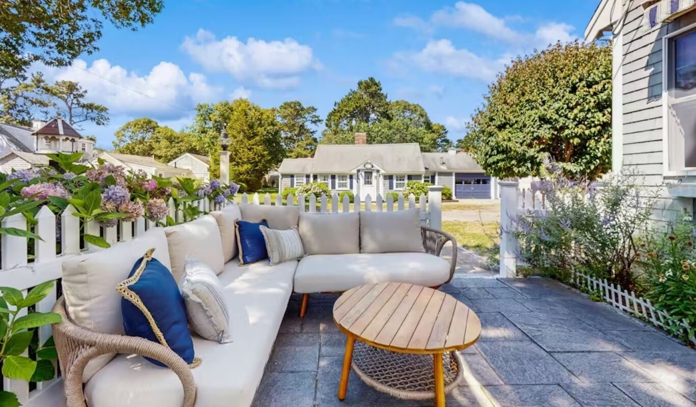 A patio with a white sofa, a round wooden table, and a white picket fence.