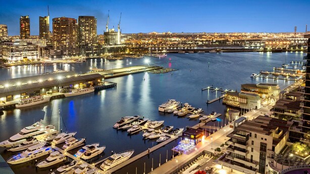 A marina with multiple boats docked, a city skyline in the background, and modern buildings with illuminated windows.